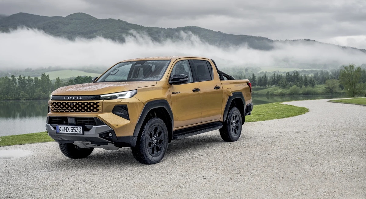 Nova Toyota Hilux Hybrid 48V amarelo mostarda (Sulphur Yellow) parada em três quartos dianteiro numa estrada de cascalho, com montanhas e neblina ao fundo