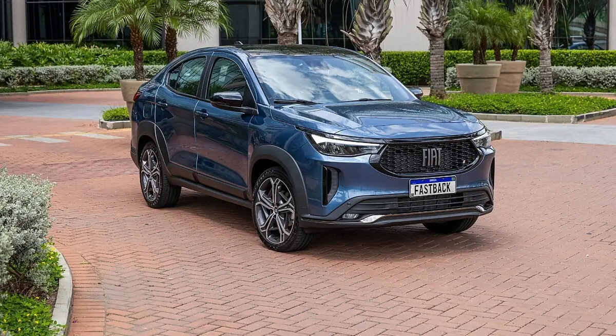 Fiat Fastback Turbo 200 Hybrid azul em vista frontal de três quartos, estacionado em pátio com palmeiras e placa de divulgação escrita FASTBACK