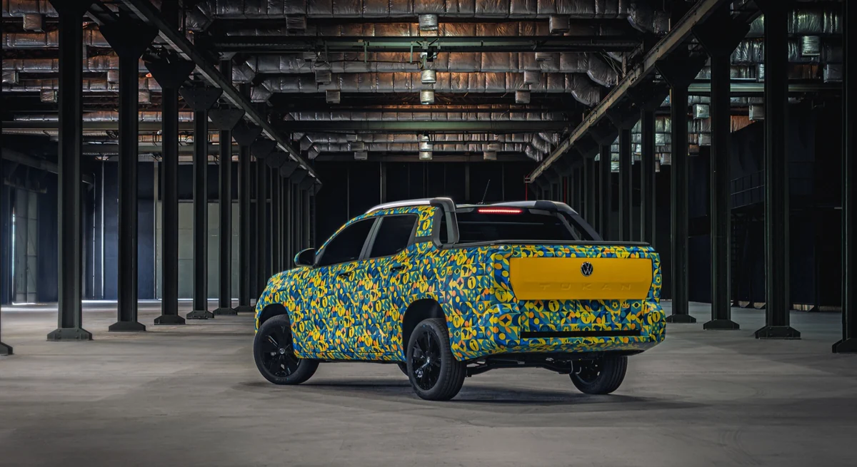 Protótipo camuflado da picape Volkswagen Tukan visto de traseira em três quartos, com tampa da caçamba amarela e nome Tukan estampado, em galpão industrial